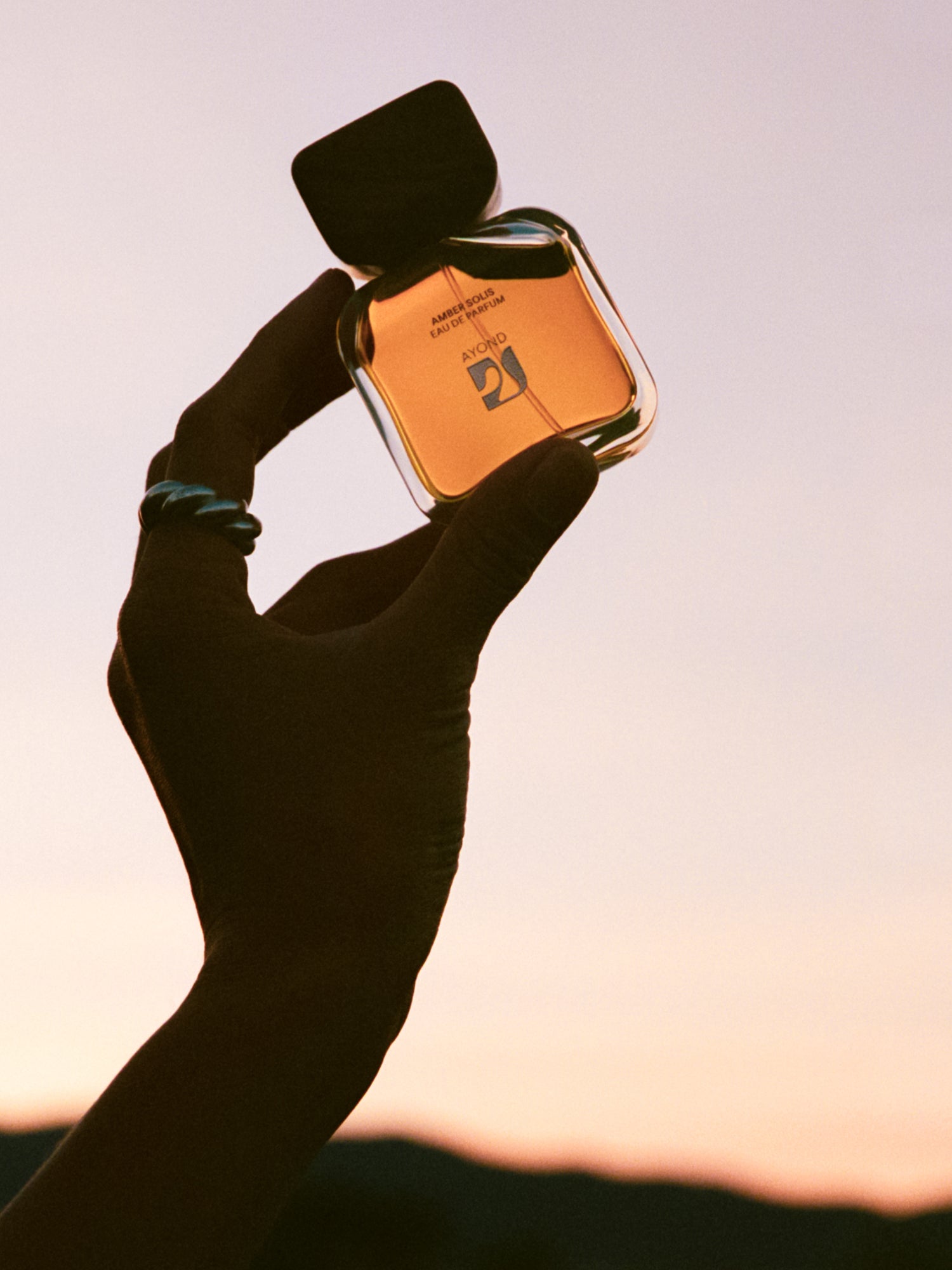 Hand holding Amber Solis Eau de Parfum bottle against a sunset sky